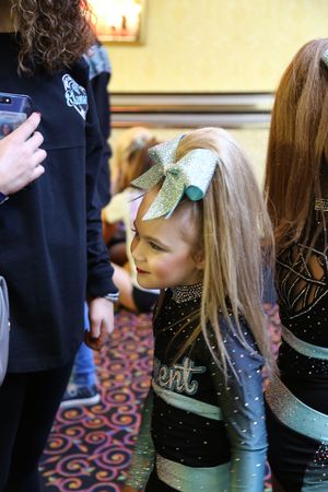 Young cheerleader with a large sparkly teal bow and rhinestone uniform looks to the side while waiting backstage at a competition.