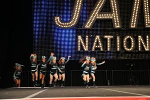 A cheer team in black and teal uniforms performs a routine on stage beneath a lit JAMZ National sign at a cheerleading competition.