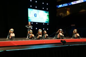 Cheerleaders in teal and black uniforms crouch in formation on a red stage at a JAMZ cheer competition with a glowing screen behind.