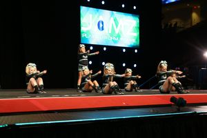 Youth cheer team performs a synchronized crouching arm motion on stage at a JAMZ cheerleading competition under bright lights.