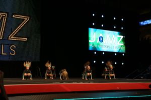 Small cheer team in black uniforms with teal accents crouches on a competition stage in front of illuminated JAMZ signs.