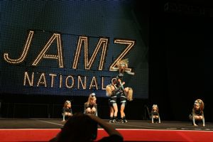 Cheer team performs a stunt on stage in front of a lit JAMZ National sign as an audience member's head appears in the foreground.
