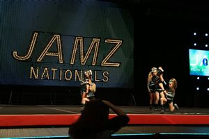 Cheer athletes perform a routine on a competition stage beneath the glowing JAMZ Nationals sign while a spectator watches.