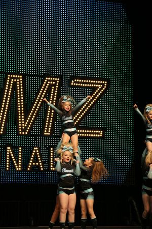 Young cheerleader in a black and teal uniform strikes a pose atop a stunt group during a JAMZ Nationals cheer performance.