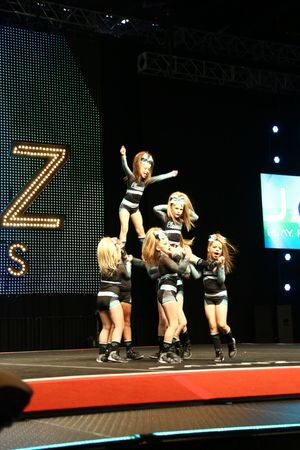 Youth cheerleaders in black and gray uniforms perform a stunt on stage with one flyer lifted high above the team.