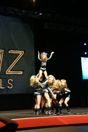 A young cheerleader stands atop a group stunt with arms raised during a competition routine on a lit stage.