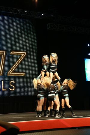Two young flyers are lifted in a double-level cheer pyramid by teammates on a competition stage with a lighted backdrop.