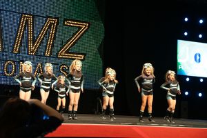 A youth cheerleading team stands in formation on a competition stage in front of a large lighted sign and video screen.