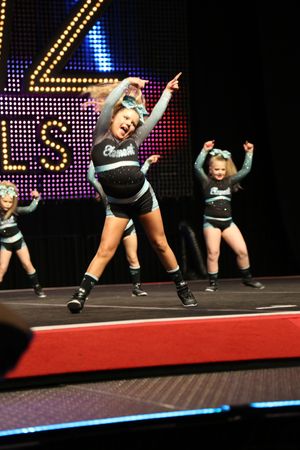 A smiling young cheerleader strikes a dynamic pose mid-routine while teammates perform behind her on a competition stage.