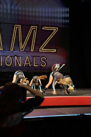Young cheerleaders bend low in a synchronized dance move on stage as a spectator films the competition in the foreground.