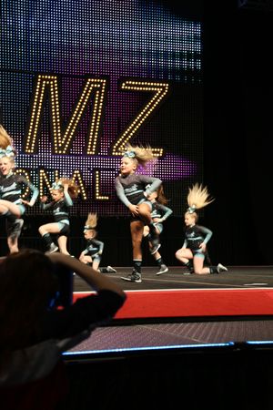 Cheerleaders leap and crouch with ponytails flying during a high-energy routine while a spectator records from the foreground.