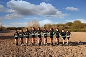 Young cheerleaders with their arms around each other face away from the camera across a sandy field beneath white clouds.