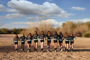 Cheer team in black and teal uniforms stands in a row facing a desert landscape under a cloudy blue sky.