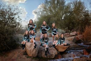 Eight young cheerleaders in matching uniforms pose on desert rocks beside a small stream during an outdoor team photo shoot.