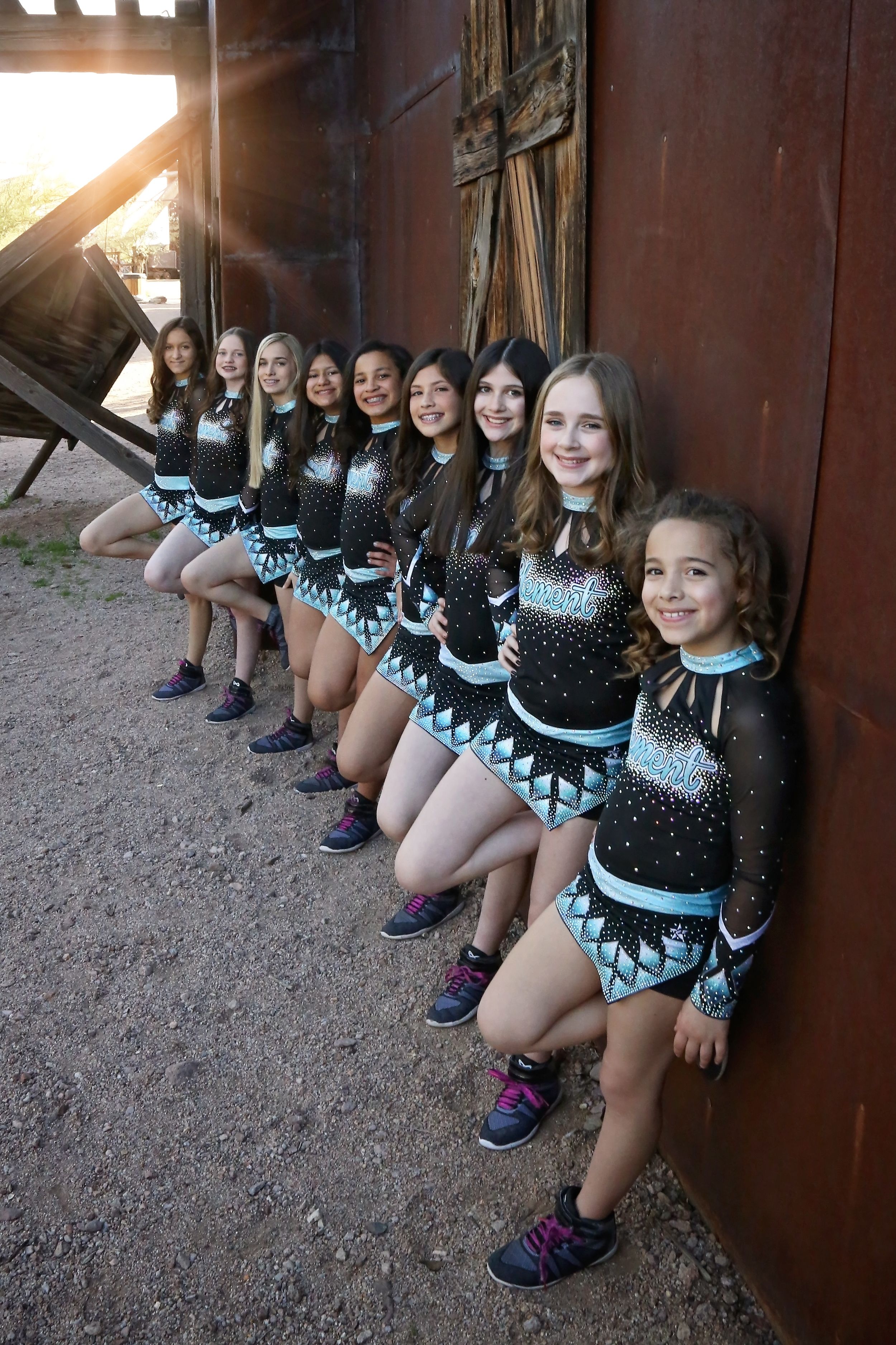 Row of smiling young cheerleaders in sparkly black and teal uniforms leaning against a rustic wall in warm golden sunlight.