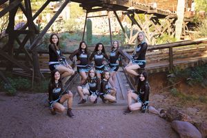 Cheer team in black and teal uniforms posing on a wooden bridge and rocks in a rustic old western town setting.