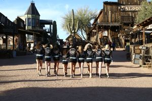 Cheerleaders walking away from the camera down a dirt street toward a western town with a saloon and tall saguaro cactus.