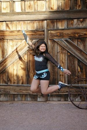 Cheerleader in a black and teal sparkly uniform leaping joyfully in front of a rustic wooden barn door beside a wagon wheel.