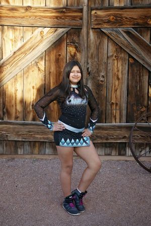 Young cheerleader in a black and teal sparkly uniform poses with hands on hips in front of a rustic wooden barn door.
