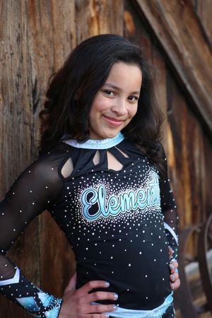 Smiling cheerleader with long curly hair wears a black Elements cheer uniform decorated with sparkling rhinestones.