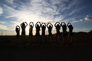 Cheerleaders raise their arms overhead in silhouette against a golden sunset and blue sky with wispy clouds.