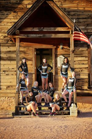 Cheer team in black and teal uniforms poses on the wooden steps of a rustic building with an American flag.