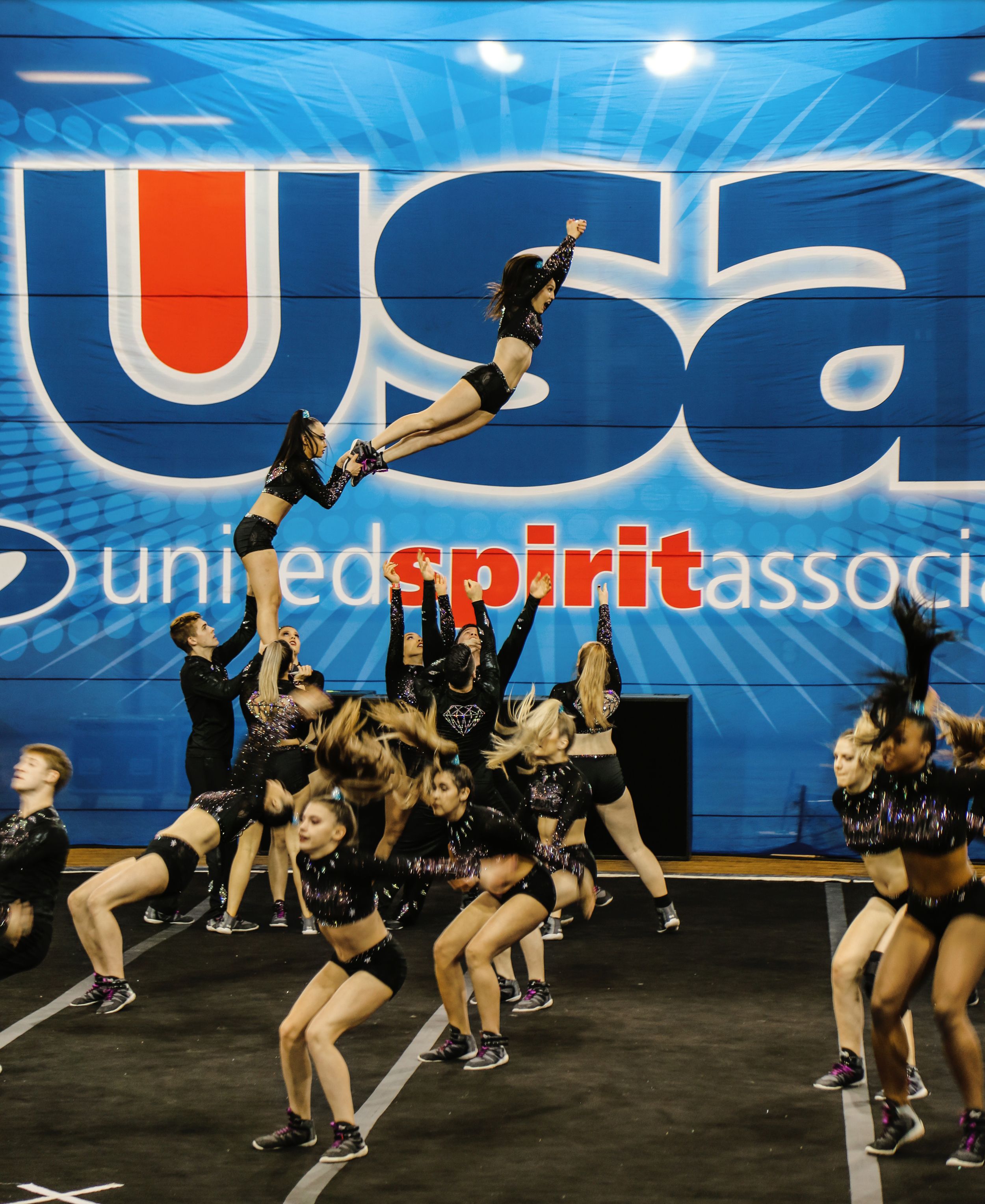 A flyer soars in a high aerial stunt above a cheer team performing on stage in front of a large USA United Spirit Association banner.