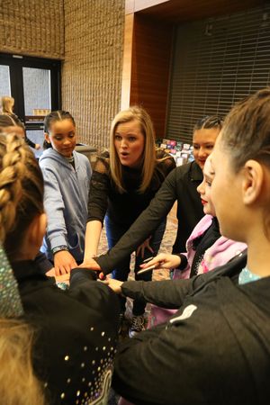 A coach and young cheer athletes put their hands together in a team huddle indoors before a competition.