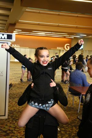 A smiling young cheerleader in a black uniform sits on a teammate's shoulders with her arms raised in an indoor convention hall.