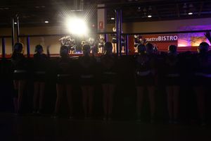 A row of cheerleaders stands in silhouette facing a brightly lit stage as they wait for their turn to perform.