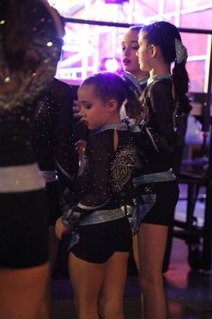 Young cheerleaders in glittery black uniforms wait backstage, with one girl looking down while teammates stand nearby.