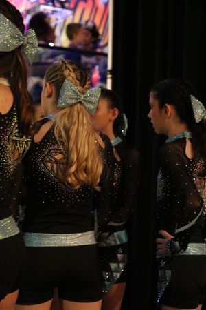 Cheerleaders in sparkly black uniforms and glittery silver bows chat together backstage at a competition.