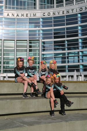 Six young cheerleaders in matching uniforms and colorful hair bows sit on stone steps in front of the Anaheim Convention Center.