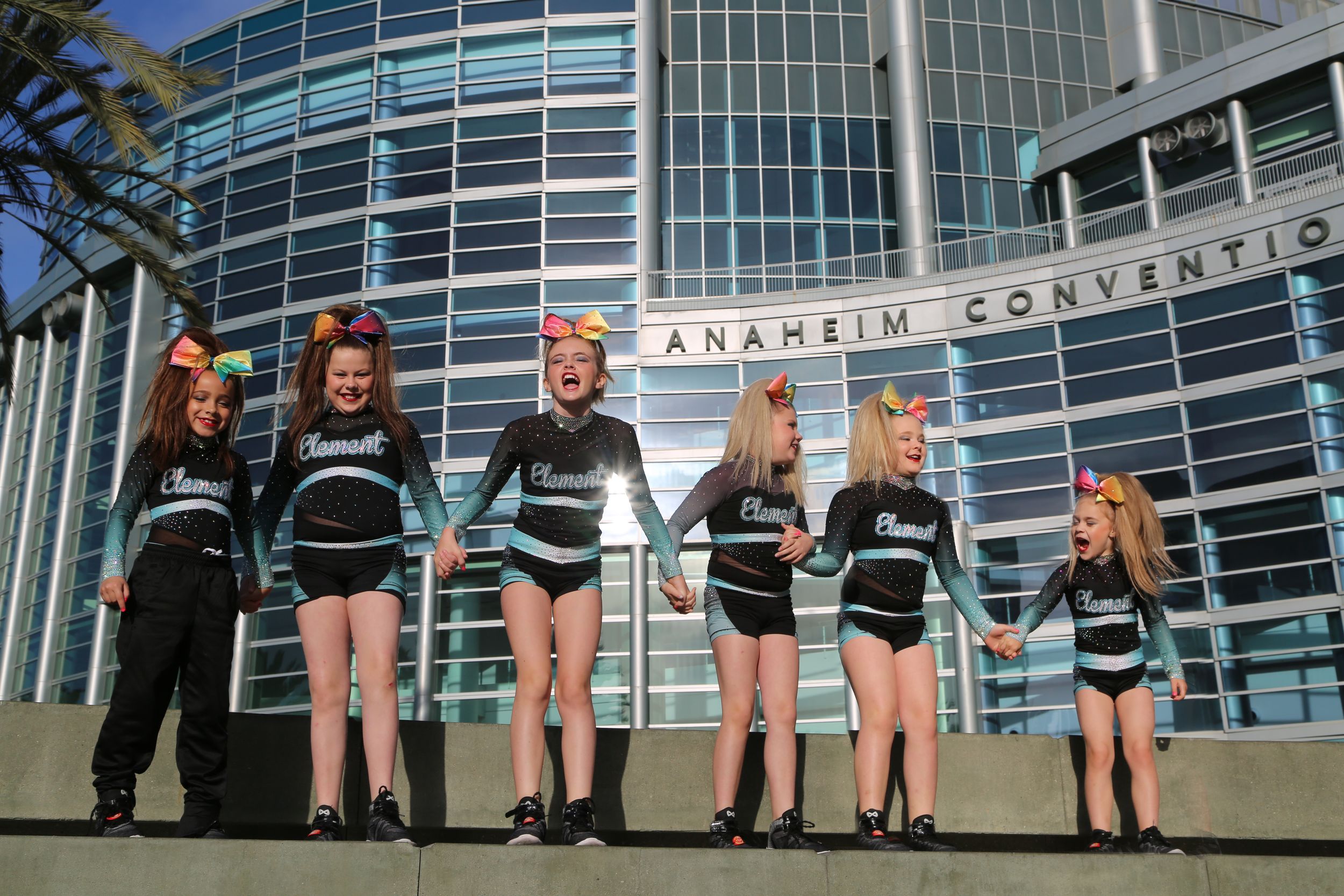 Young cheerleaders in matching black and teal uniforms hold hands in a line outside the Anaheim Convention Center building.