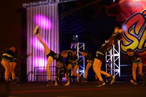 Cheerleaders in black uniforms perform a synchronized high kick routine on stage under bright purple lights.