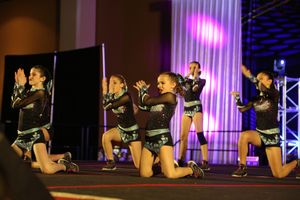 Young cheer team in sparkly black uniforms kneels in formation on the competition stage during a dance routine.