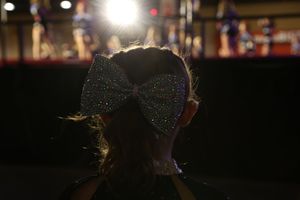 A girl wearing a large sparkling rhinestone bow is silhouetted against bright stage lights at a cheerleading competition.