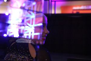 A smiling cheerleader in a sparkly uniform is lit by colorful purple and blue lights backstage at a competition.