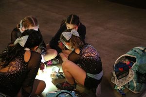 Cheerleaders with glittery bows sit in a circle on the floor, huddled over a small light and their gear bags.