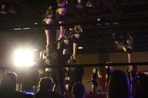 Cheerleaders perform a stunt sequence with flyers lifted high in the air under a warehouse-style ceiling at a competition.
