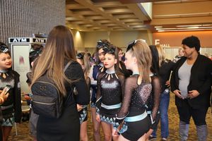 Cheerleaders in black sparkly uniforms gather in a convention hall hallway, talking with parents and coaches before competing.