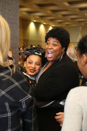 A laughing woman in a black blazer embraces a smiling cheerleader wearing a bow and red lipstick at a competition.