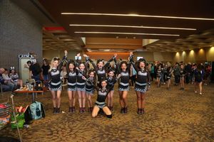 A cheer team in black uniforms poses with arms raised in a convention center hallway as onlookers watch nearby.