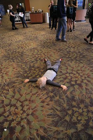 A young child in gray clothing lies spread-eagle on a patterned carpet while adults stand and talk in the background.