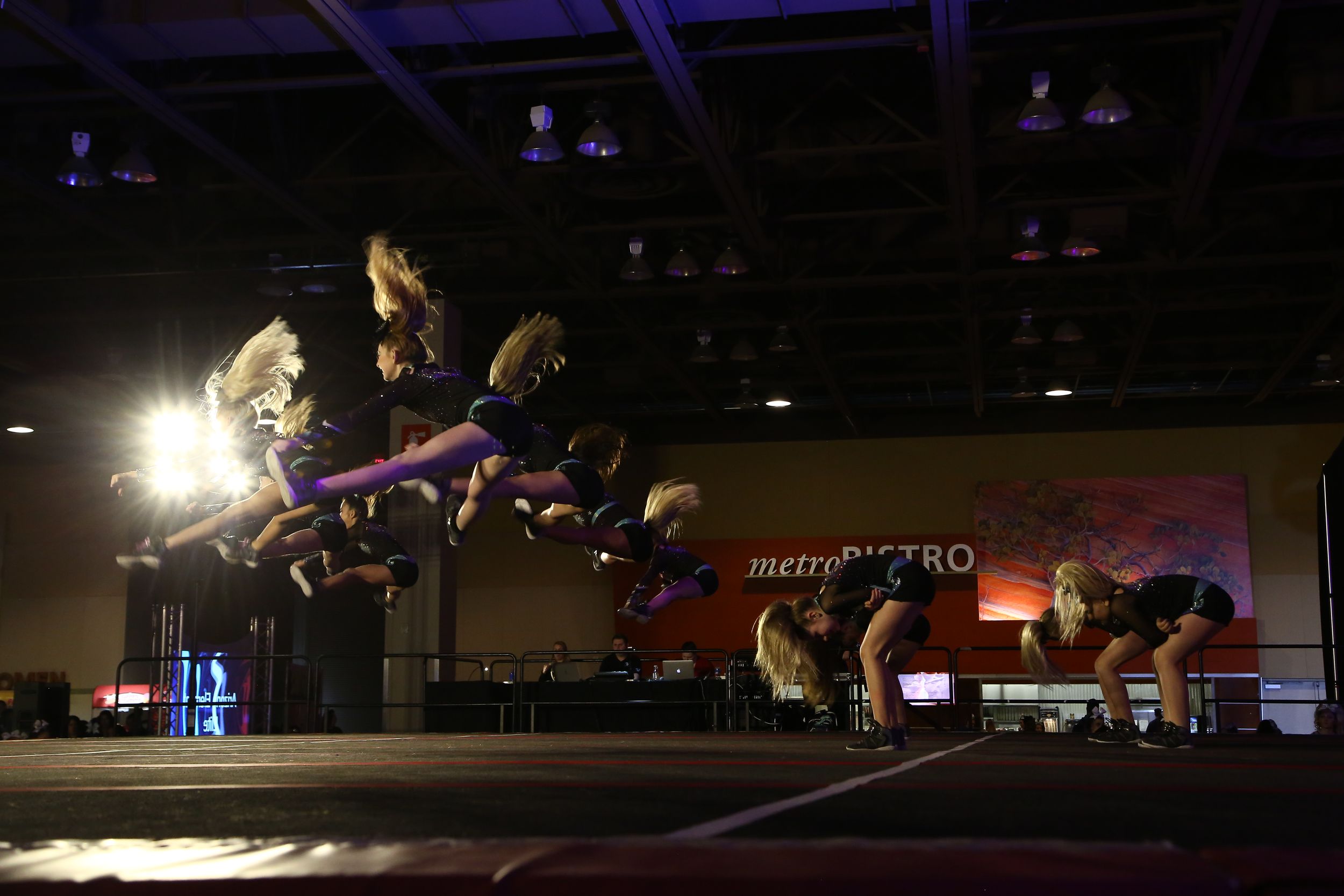 A cheerleading squad performs a tumbling and stunt routine on a competition stage under bright lights, captured in motion blur.