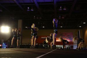 Cheerleaders in black uniforms perform a synchronized routine on a competition stage beneath bright stage lights.