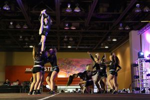 Cheerleaders lift a flyer high into the air during a stunt on a competition stage in a large arena.