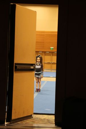 A young cheerleader in a sparkly uniform and hair bow stands in a doorway, waiting to take the competition floor.