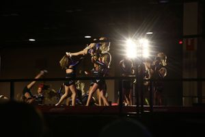 Cheerleaders perform a stunt on a dark competition stage as bright spotlights glow behind the team.