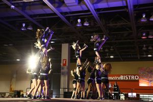 A cheer team lifts several flyers into the air in a pyramid stunt on a competition stage in a large arena.